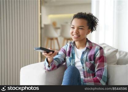 Attractive teenage girl is watching it and shifts channels with remote controller. Reality show or series watching. Positive african american or hispanic young woman has weekend leisure at home.. Attractive teenage girl is watching it and shifts channels with remote controller. Weekend leisure.