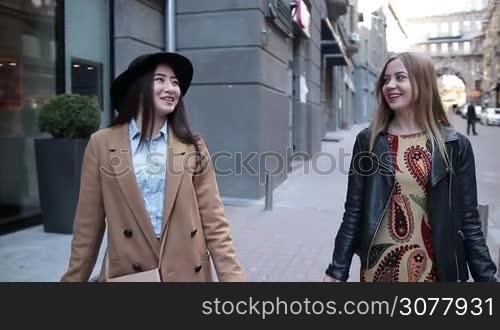 Attractive teenage girlfriends walking with shopping bags on the street, looking happy. Stylish asian woman in black hat chatting with pretty caucasian female friend while strolling down the sidewalk after shopping. Slow motion.