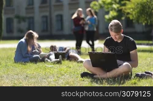 Attractive teenage hipster sitting on green grass at campus lawn while studying outdoors. Handsome university student using laptop computer while doing homework on park lawn with blurry group of students studying and relaxing on background.