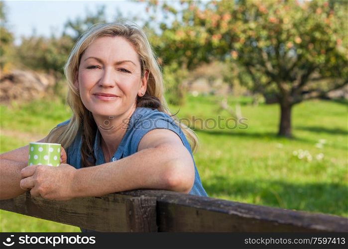Attractive thoughtful middle aged woman leaning resting on fence in the countryside with mug or cup of tea or coffee