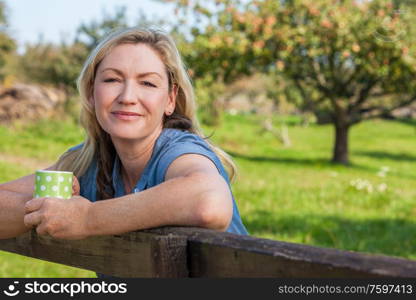 Attractive thoughtful middle aged woman leaning resting on fence in the countryside with mug or cup of tea or coffee