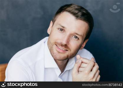 Attractive unshaven male office worker has break after work, looks joyfully at camera, going to have good rest, wears elegant white shirt, poses indoor alone. People, style and occupation concept