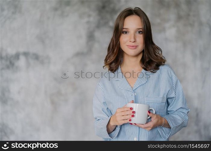 Attractive woman dressed in nightwear, holds white mug with coffee or tea, has morning drink, poses indoor against blurred background with copy space for your advertisement or promotional content