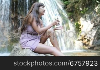 attractive woman drinking water from plastic bottle by the waterfall