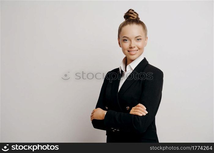 Attractive woman executive in dark suit with hair in bun, standing sideways looking forward with her arms crossed radiating calmness and confidence isolated on grey background. Business people concept. Woman executive in dark suit standing sideways