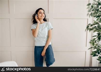 Attractive woman in headphones standing at the wall and listen to music. Pretty lady in earphones relax in the room, female sound lover resting. Attractive woman in headphones listen to music