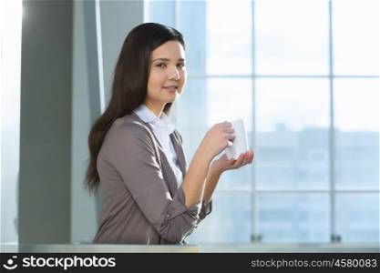 Attractive woman in office building. Young brunette woman in modern glass interior with mug in hands