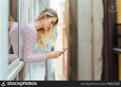 Attractive woman leaning out of her house window using a smart phone.. Attractive woman leaning out of her house window using a smartphone.