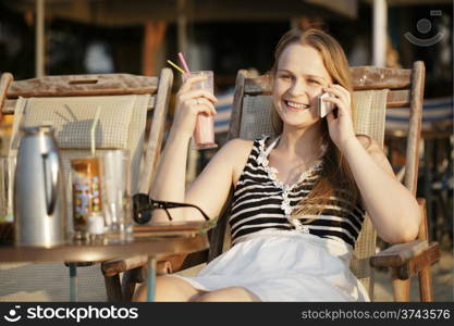 Attractive woman relaxing in a deckchair in the sun talking on her mobile phone and drinking cocktail. Horizontal shot.