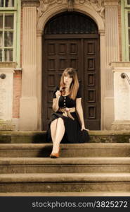 Attractive woman sitting on steps outdoors. Retro style fashionable girl in black classic dress and sunglasses relaxing on street of the old town city Gdansk Danzig Poland