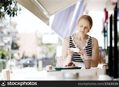 Attractive woman taking picture of a pastry on her smart phone as she sits at a table at an open-air restaurant enjoying refreshments