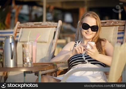 Attractive woman wearing sunglasses relaxing in a deckchair in the sun sending an sms on her mobile phone