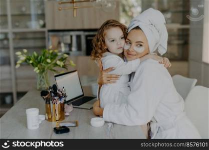 Attractive woman wears hydrogel patches under eyes dressed in bathrobe and wrapped towel embraces her small daughter with love pose together at home near cosmetic tools. Makeup routine concept