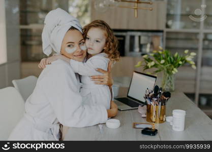 Attractive woman wears hydrogel patches under eyes dressed in bathrobe and wrapped towel embraces her small daughter with love pose together at home near cosmetic tools. Makeup routine concept