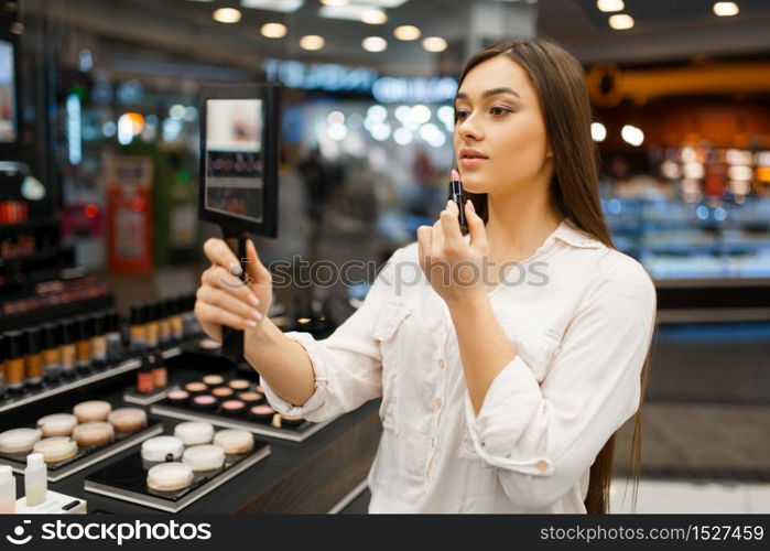 Attractive woman with mirror applies lipstick in cosmetics store. Buyer at the showcase in luxury beauty shop salon, female customer in fashion market. Woman with mirror applies lipstick in store