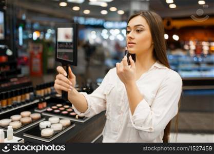Attractive woman with mirror applies lipstick in cosmetics store. Buyer at the showcase in luxury beauty shop salon, female customer in fashion market. Woman with mirror applies lipstick in store