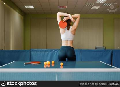 Attractive woman with racket poses at the ping pong table indoors, back view. Female person in sportswear, training in table tennis-club