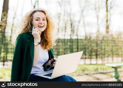 Attractive woman with trendy hairdo sitting in park communicating over her cell phone using modern laptop computer having broad smile while looking aside. People, technology, communication concept