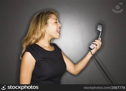 Attractive woman yelling into phone receiver standing against gray background.