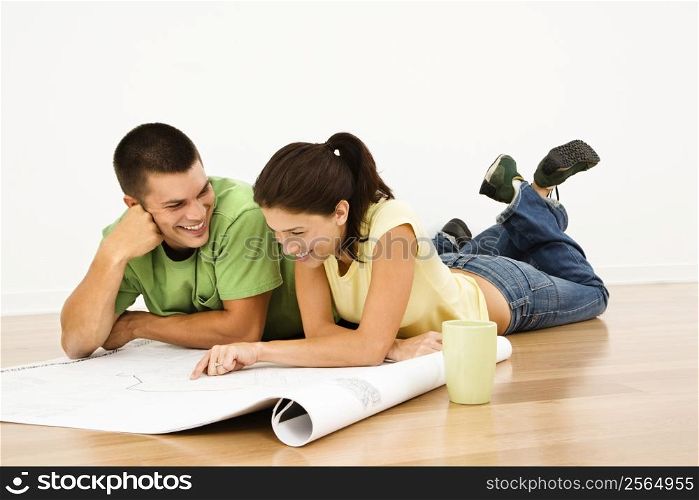 Attractive young adult couple lying on home floor with coffee cups smiling and looking at blueprints.