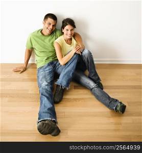 Attractive young adult couple sitting close on hardwood floor in home smiling.