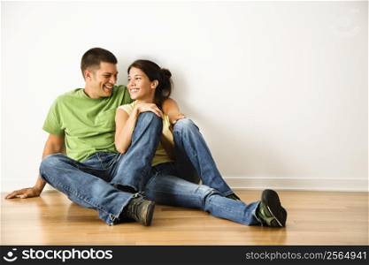 Attractive young adult couple sitting close on hardwood floor in home smiling.