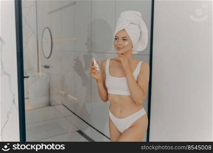 Attractive young adult woman applying facial cream, beautiful healthy lady with head wrapped in towel putting moisturizing lifting nourishing day cream on clean soft skin in bathroom at home. Attractive young adult woman applying facial cream after morning shower