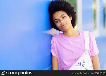 Attractive young African American female in casual clothes looking at camera while leaning on blue building on street of city. Charming black woman near blue house
