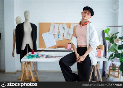 Attractive young Asian LGBTQ gay dressmaker sitting pose on working desk, confident looking at camera, sketch of fashion dress design and mannequin in studio, small tailor business owner concept