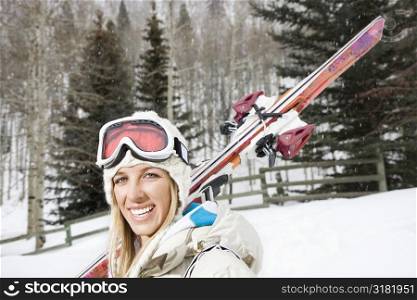 Attractive young blond woman in winter ski gear carrying skis over shoulder smiling.