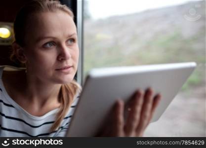 Attractive young blond woman traveling on a train with a tablet held in her hand as she stares thoughtfully into the air