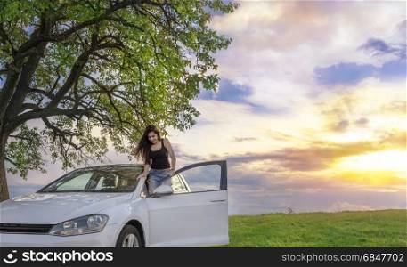 Attractive young brunette enjoys her car, in the spring nature as the sun is setting down.