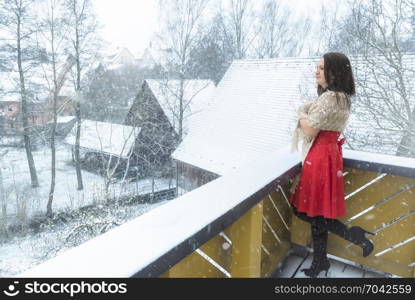 Attractive young brunette woman, in a red dress and black boots, covered with a handmade shawl, stands on the balcony and enjoys the first snow.