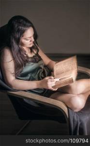Attractive young brunette woman, with long hair, dressed sexy, sitting in a cozy armchair and reading from an old book.