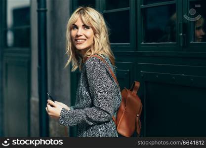Attractive young caucasian woman looking at her smart phone in urban background. Blonde girl wearing casual clothes outdoors