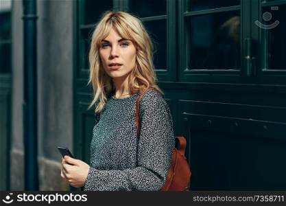 Attractive young caucasian woman looking at her smart phone in urban background. Blonde girl wearing casual clothes outdoors. Attractive young caucasian woman looking at her smart phone outdoors