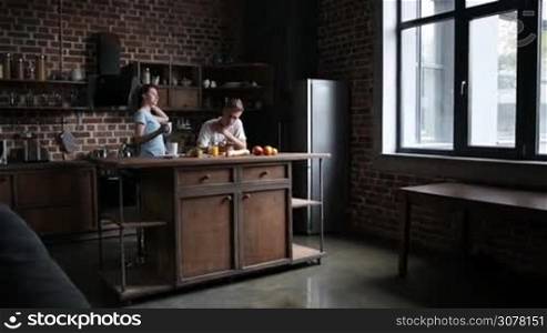 Attractive young couple bonding and enjoying leisure in the kitchen in the morning. Beautiful girl bringing cup of hot coffee to her boyfriend while man surfing the net on smartphone. Slow motion. Steadicam stabilized shot.