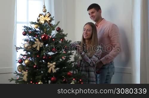 Attractive young couple in love decorating Christmas tree with colorful glass baubles at home. Smiling man kissing his beautiful wife and embracing her from behind while carefree family putting decorative balls on xmas fir tree in living room.