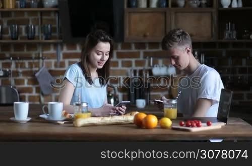 Attractive young couple sitting at kitchen table and surfing the net with smartphones while having breakfast together. Cheerful hipsters socializing through mobile phones and sharing latest gossips and news while spending lovely morning at home.