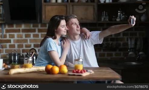 Attractive young couple taking selfie with smart phone and smiling while sitting at the kitchen table and having breakfast in the morning. Cheerful family spending leisure together at home and making selfie with mobile phone.