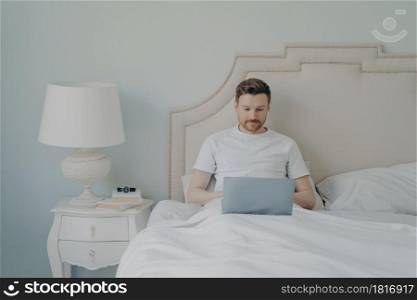 Attractive young european man watching video online on portable laptop computer while sitting in comfortable bed, using free internet connection at home. Leisure time indoor concept. Attractive young european man watches video online on portable laptop computer while lying in bed