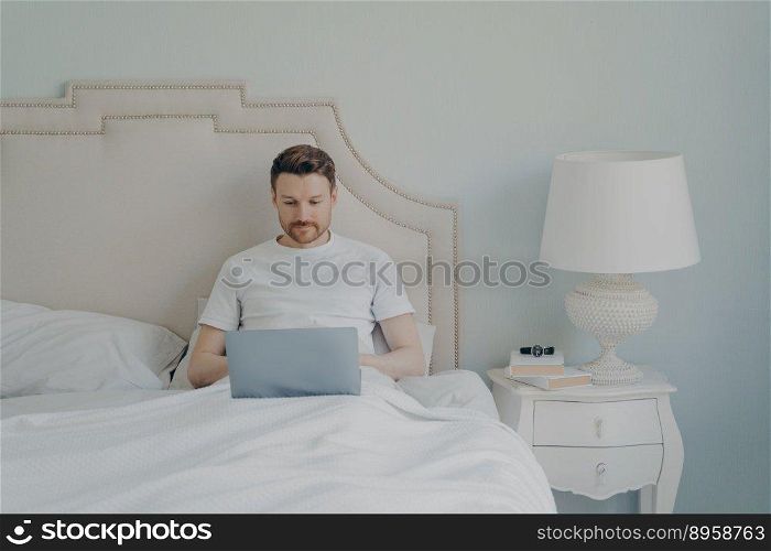 Attractive young european man watching video online on portable laptop computer while sitting in comfortable bed, using free internet connection at home. Leisure time indoor concept. Attractive young european man watches video online on portable laptop computer while lying in bed