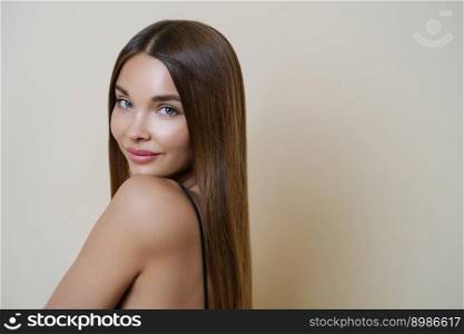 Attractive young European woman with soft healthy skin, perfect body, looks calm directly at camera, has long hair, stands indoor against beige background. Youth, beauty and skin care concept.