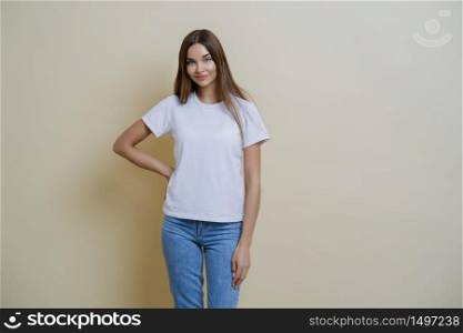 Attractive young female keeps hand on waist, wears casual clothes, ready for going out during weekend, enjoys nice weather, expresses joyful positive attitude to interlocutor, poses over beige wall