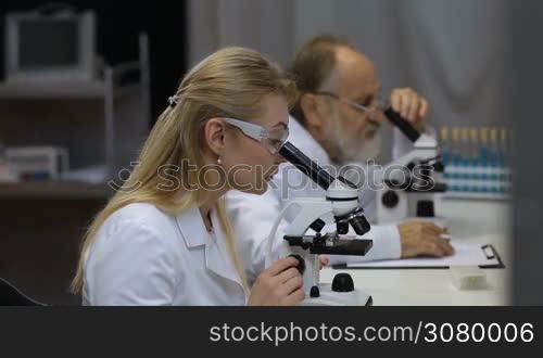 Attractive young female scientist and her senior male supervisor pipetting and microscoping in the life science research laboratory. Young female research scientist and senior male supervisor preparing and analyzing microscope slides in research lab.