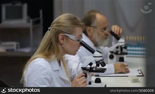 Attractive young female scientist and her senior male supervisor pipetting and microscoping in the life science research laboratory. Young female research scientist and senior male supervisor preparing and analyzing microscope slides in research lab.