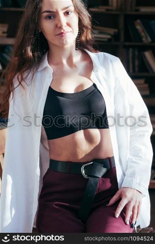 Attractive young female wearing black sport bra lingerie and white shirt unbuttoned, casual fashion style, sitting on wooden chair, bookshelves in the backdrop. Long hair, sensual beautiful seductive
