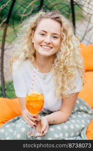 Attractive young female with charming smile, has wavy hair, dressed casually, holds fresh summer cocktail, recreats in garden during weekend. People, rest and lifestyle concept. Vertical shot