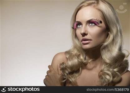 attractive young girl with long blonde wavy hair posing in beauty portrait with pink feathered, creative make-up and shiny decorations on the face