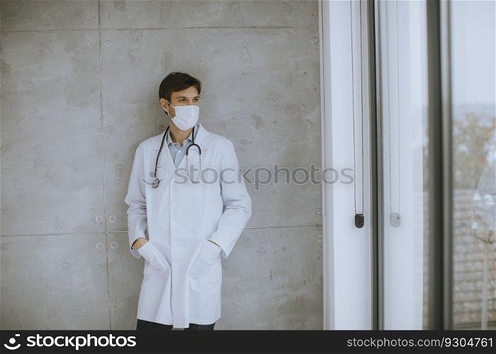 Attractive young male doctor wearing surgical protective mask and standing by the wall